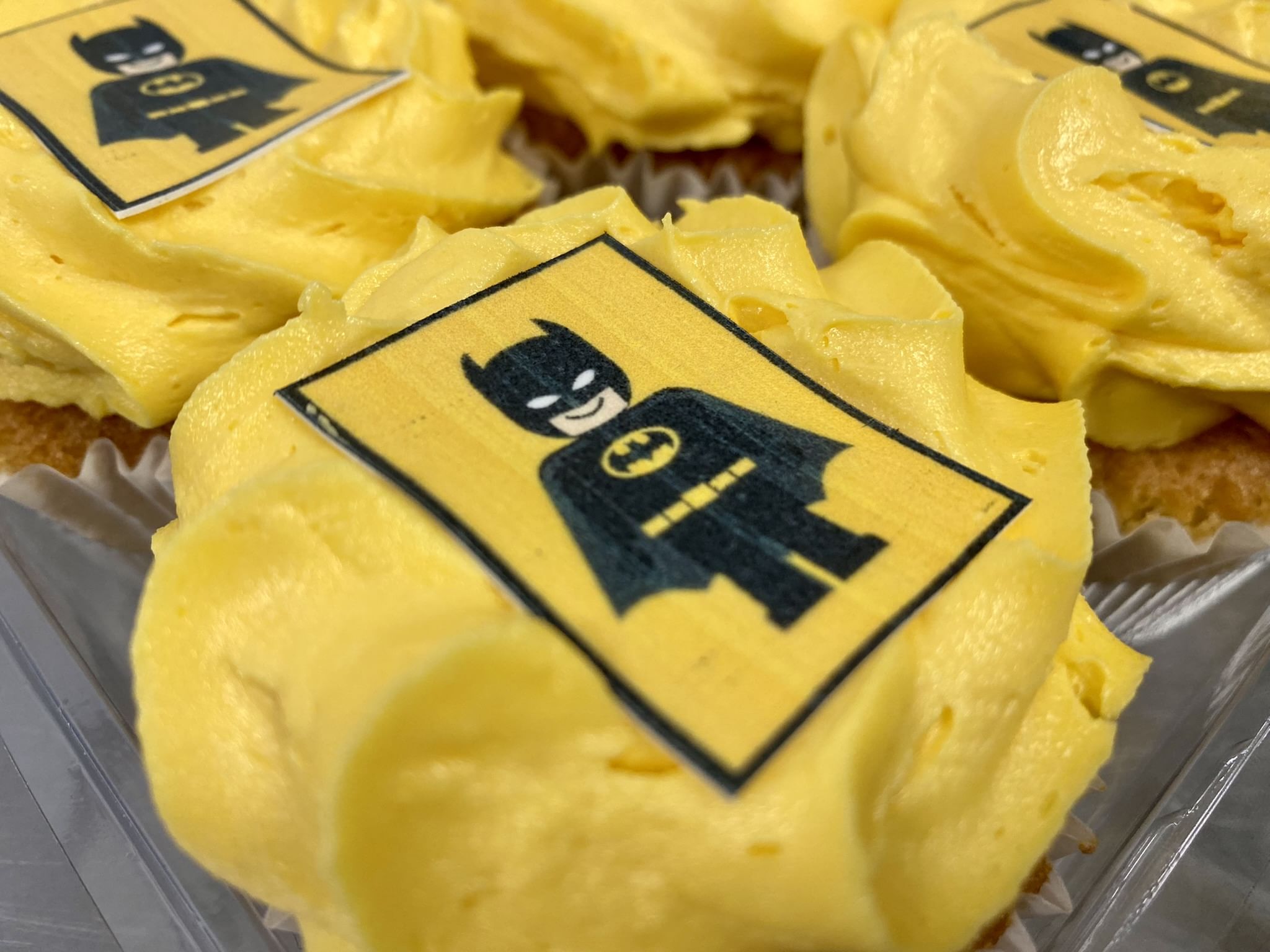 Cupcakes topped with yellow frosting and edible images of a LEGO-style Batman figure in a black superhero costume, featuring bat ears and a bat logo on the chest. These custom-printed cupcakes are arranged in a close-up view, showcasing the detailed decoration.